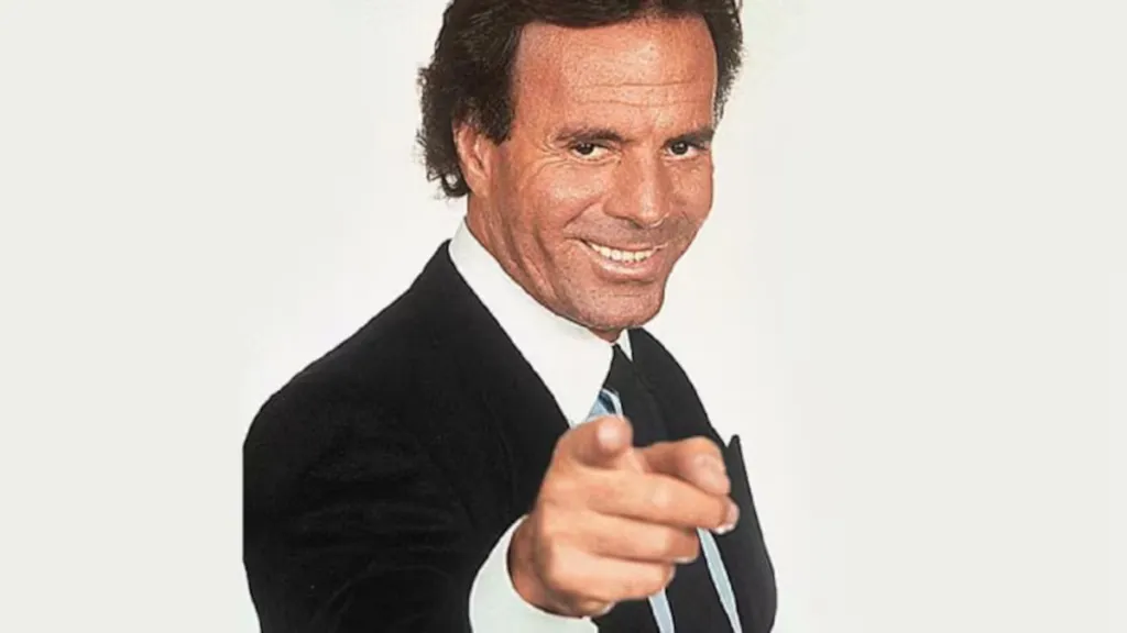 Julio Iglesias smiling in a dark suit and pointing directly at the camera with his index finger.