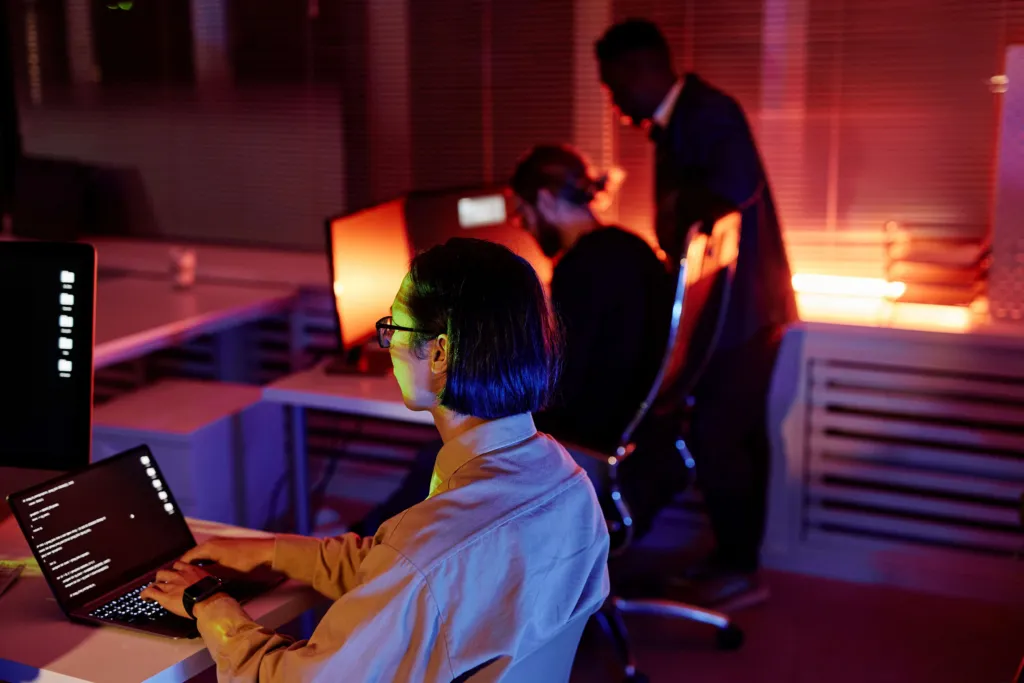 Group of colleagues working late at night in a modern office with lit screens and city lights in the background